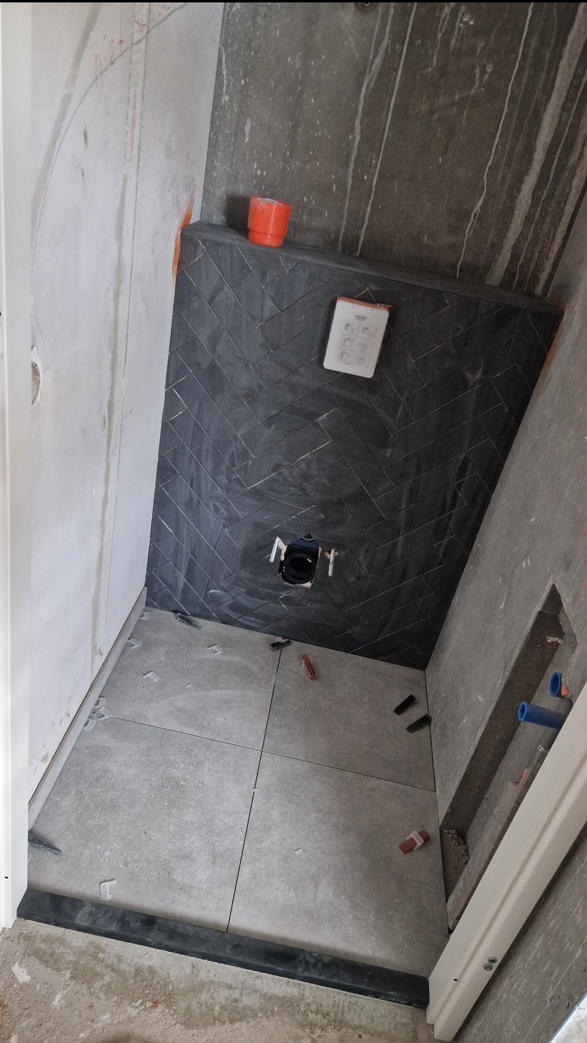 Wnętrze łazienki w trakcie prac wykończeniowych: czarne płytki ułożone w jodełkę na ścianie, szare płytki podłogowe, widoczne elementy instalacji i akcesoria montażowe.