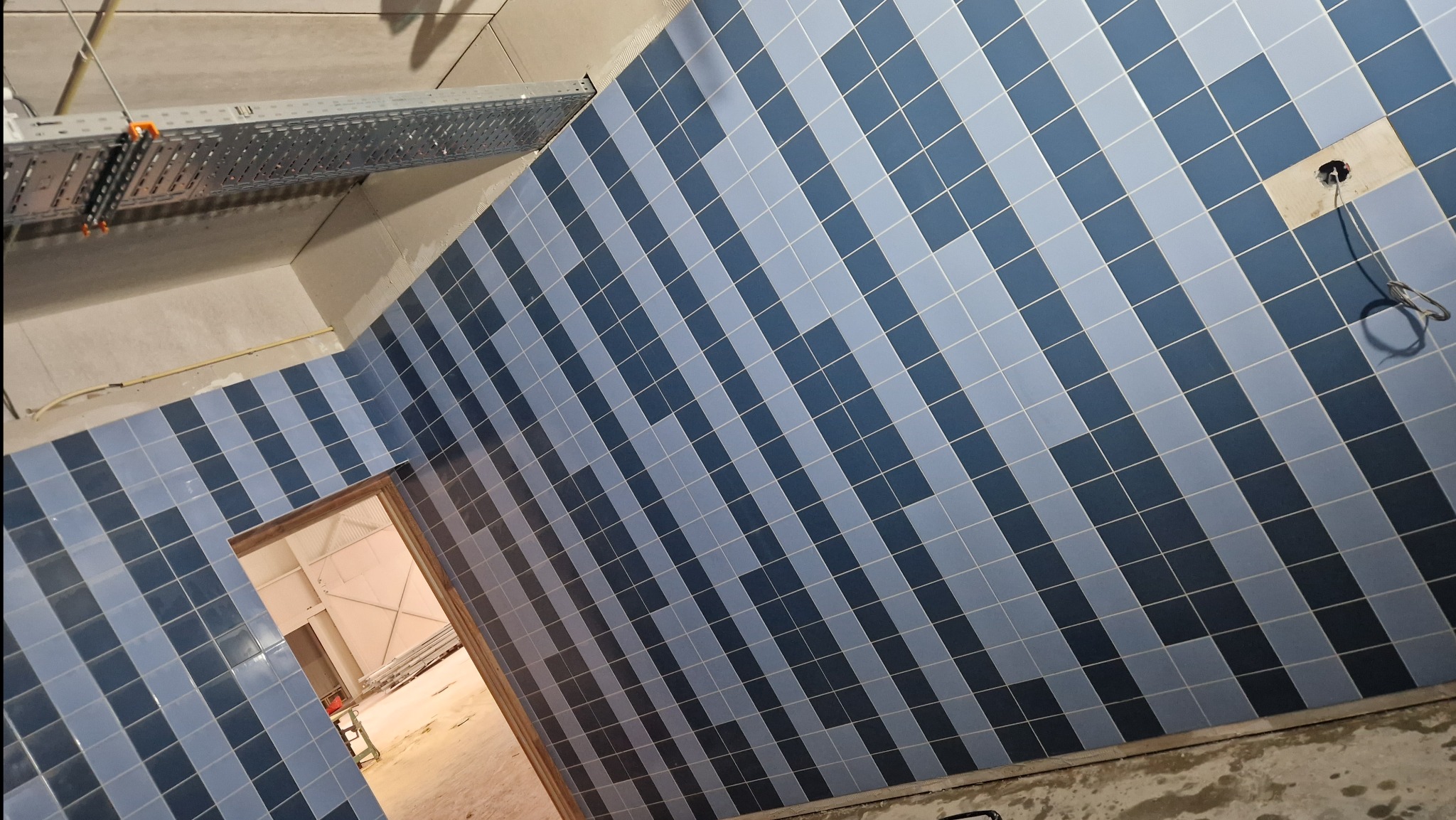 Ściana w trakcie układania niebiesko-szarej szachownicy z płytek ceramicznych, widoczne kable wystające ze ściany oraz metalowa konstrukcja pod sufitem.