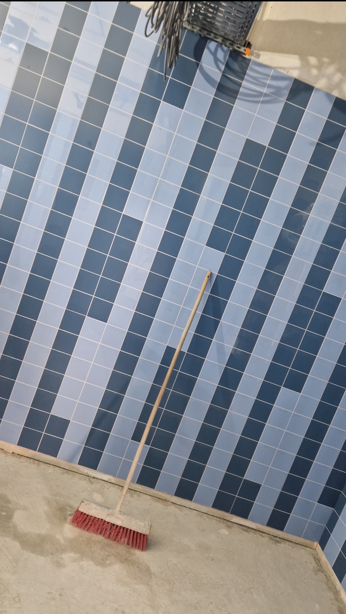 Niedokończona łazienka z niebiesko-białymi kafelkami ułożonymi ukośnie na ścianie i czerwoną zmiotką stojącą na surowej podłodze.