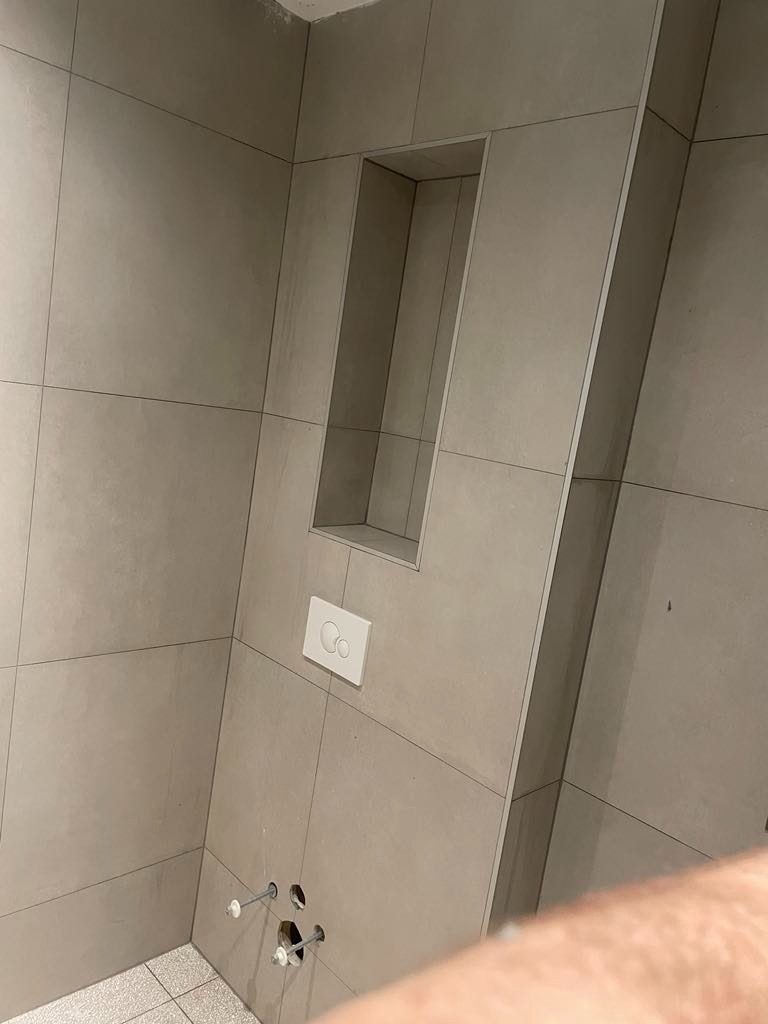 Ściana łazienki wyłożona jasnoszarymi płytkami z wnęką, widoczny przycisk spłuczki i wystające rurki instalacyjne.