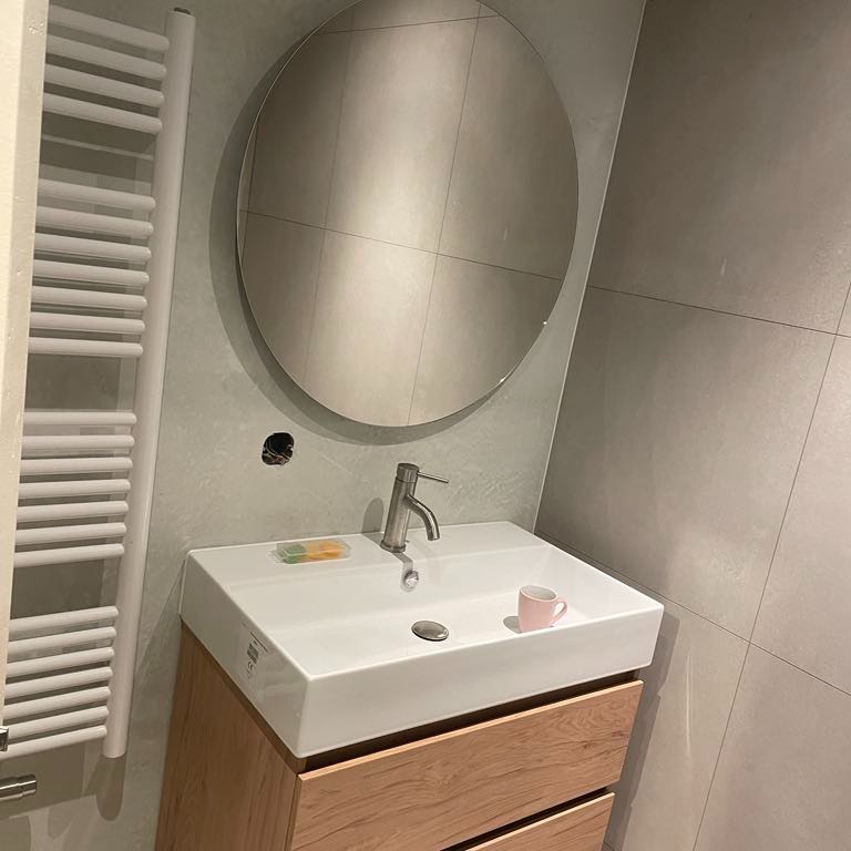 Nowoczesna łazienka z okrągłym lustrem, białą umywalką nablatową na drewnianej szafce, grzejnikiem drabinkowym i szarymi płytkami; na umywalce mała różowa filiżanka.