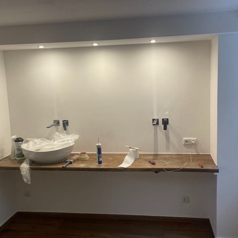 Niedokończona łazienka z drewnianym blatem, na którym stoi biała umywalka owinięta folią, obok silikon, taśma malarska i narzędzia, widoczne wystające rury do armatury i gniazdko elektryczne.