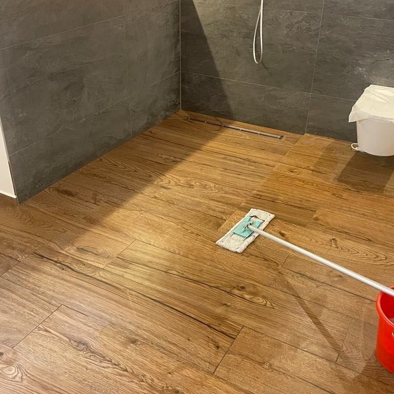 Podłoga z paneli imitujących drewno w łazience, częściowo umyta mopem płaskim; widoczna szara ściana z płytek oraz wiadro.