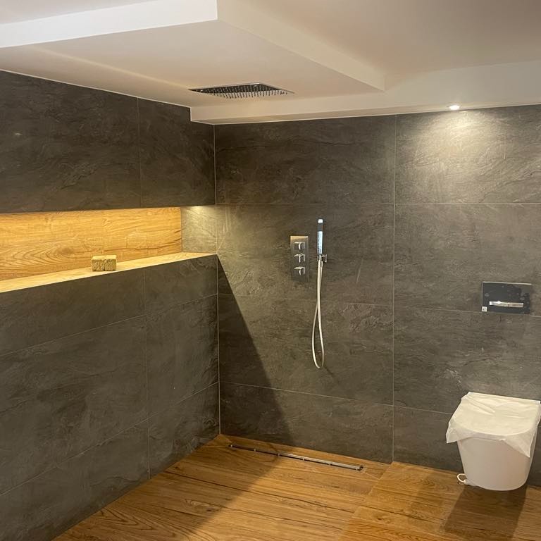 Nowoczesna łazienka z ciemnymi, matowymi płytkami imitującymi kamień na ścianach, drewnianą podłogą i wnęką z drewnianą półką. Uwagę zwraca minimalistyczna bateria prysznicowa i toaleta...
