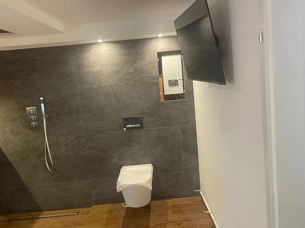 Nowoczesna łazienka z szarymi płytkami imitującymi kamień, drewnianą podłogą, zabudowanym prysznicem, podwieszaną toaletą przykrytą folią ochronną i telewizorem zamontowanym na białej ścianie.