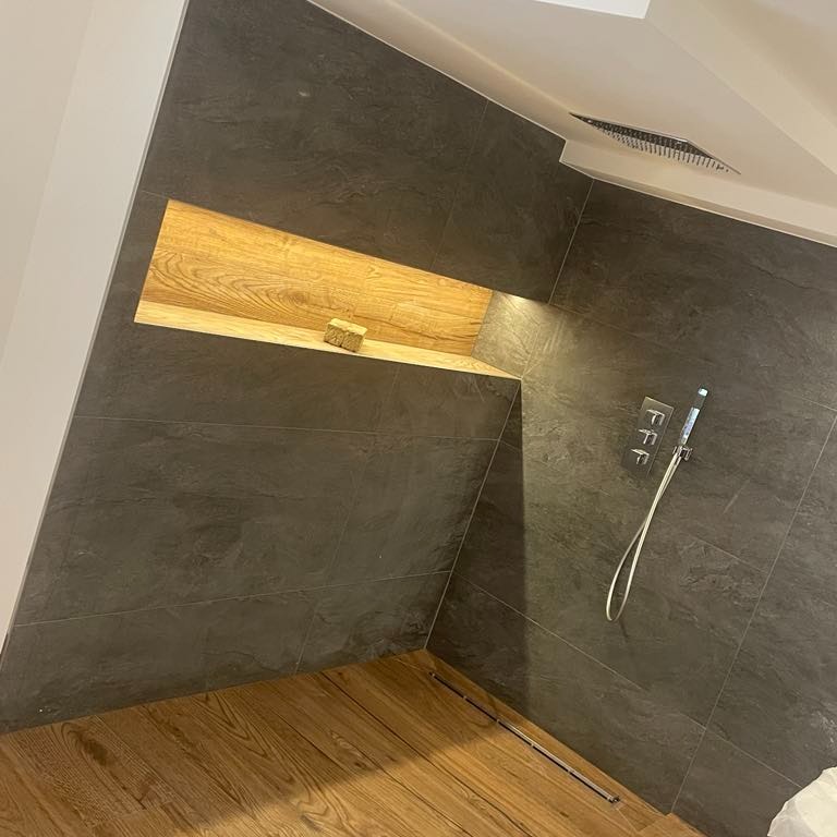 Nowoczesna kabina prysznicowa z szarymi płytkami imitującymi kamień, drewnianą półką wnękową z kostką mydła, drewnianą podłogą i chromowaną baterią prysznicową z trzema pokrętłami.