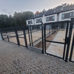 Jakub Grabowicz - Nowoczesne ogrodzenie z paneli zgrzewanych w kolorze grafitowym, zabezpieczające teren posesji z nowym budynkiem mieszkalnym w tle, widok z poziomu kostki brukowej.