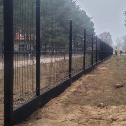 Jakub Grabowicz - Długi, czarny system ogrodzeniowy z siatki panelowej, z betonową podmurówką, w trakcie montażu w pochmurny dzień; widoczni pracownicy w oddali.