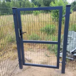 Szara furtka z zamkiem w siatce ogrodzeniowej na tle zarośniętego pola i drzew, widoczne ślady montażu, przy słupku leżą plandeki.