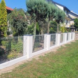 Jakub Grabowicz - Nowoczesne ogrodzenie z siatki z białymi słupkami, widok z boku, w tle zadbany ogród i domy.