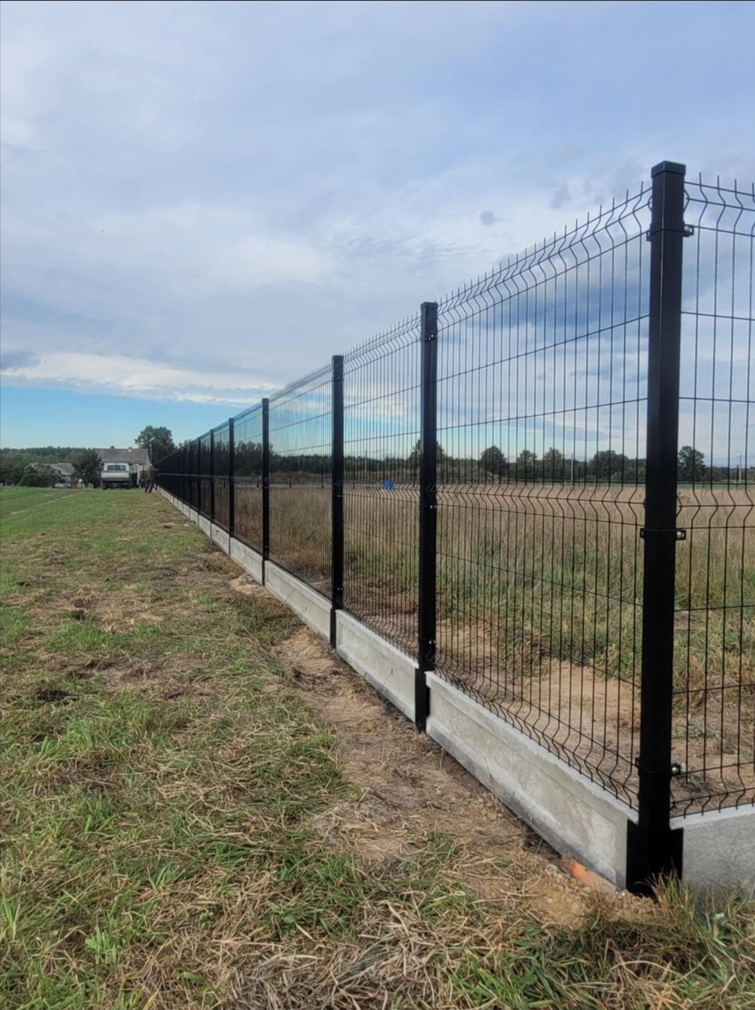 Długie, czarne ogrodzenie z paneli z podmurówką betonową, rozciągające się wzdłuż trawiastego terenu w kierunku zabudowań w tle, widoczne pod pochmurnym niebem.