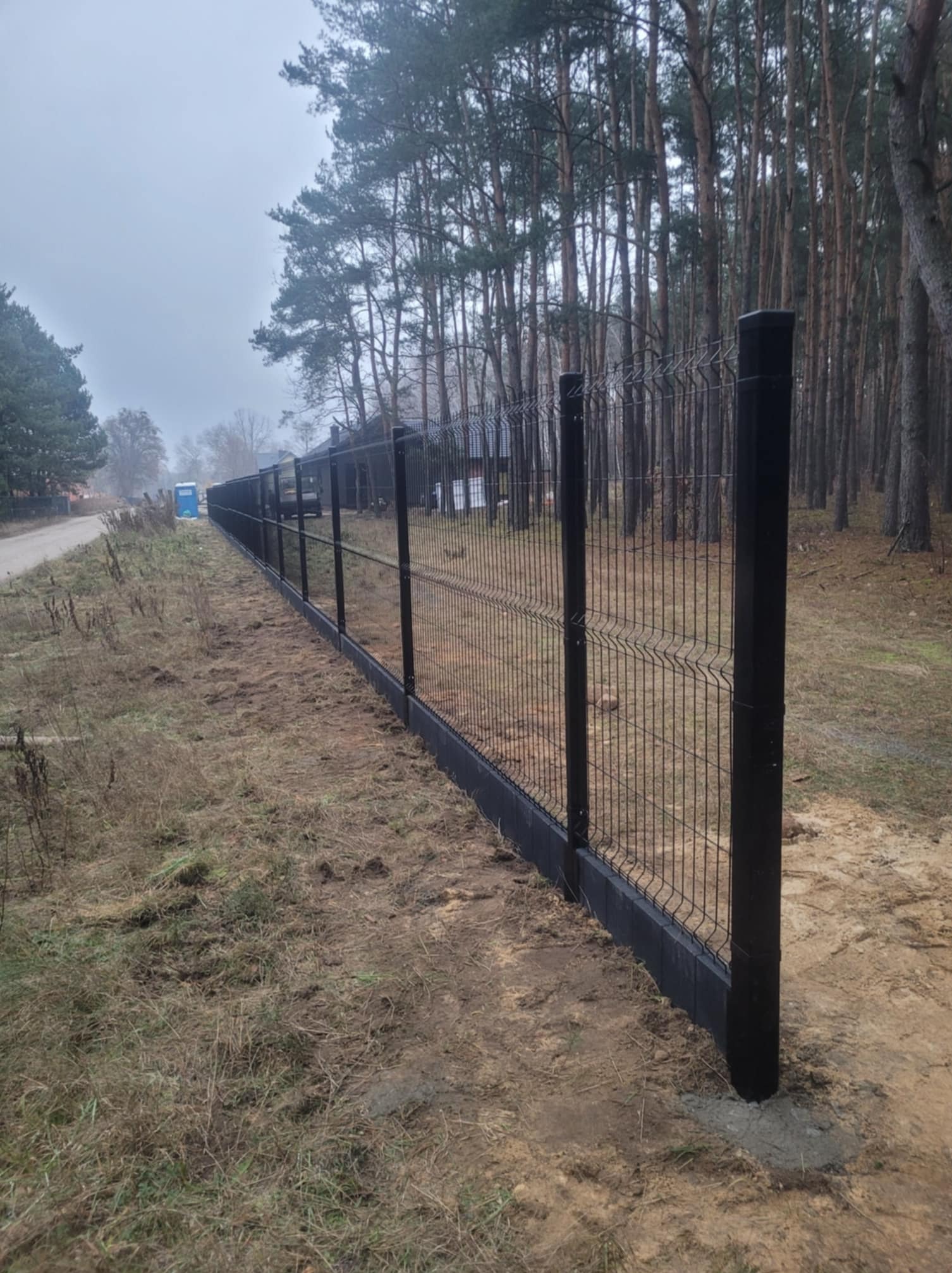 Długi, czarny panel ogrodzeniowy z podmurówką betonową, ciągnący się wzdłuż polnej drogi na tle lasu w pochmurny dzień.