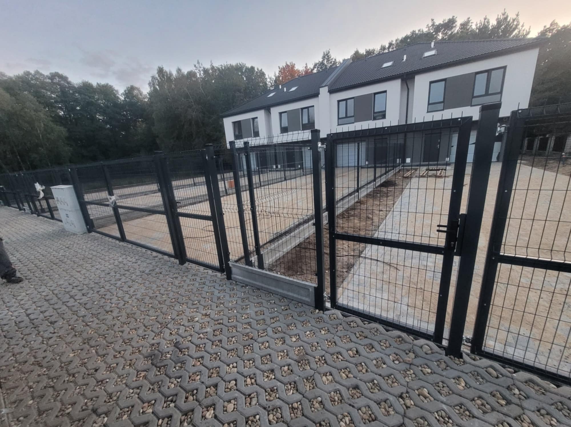 Nowoczesne ogrodzenie z paneli zgrzewanych w kolorze grafitowym, zabezpieczające teren posesji z nowym budynkiem mieszkalnym w tle, widok z poziomu kostki brukowej.
