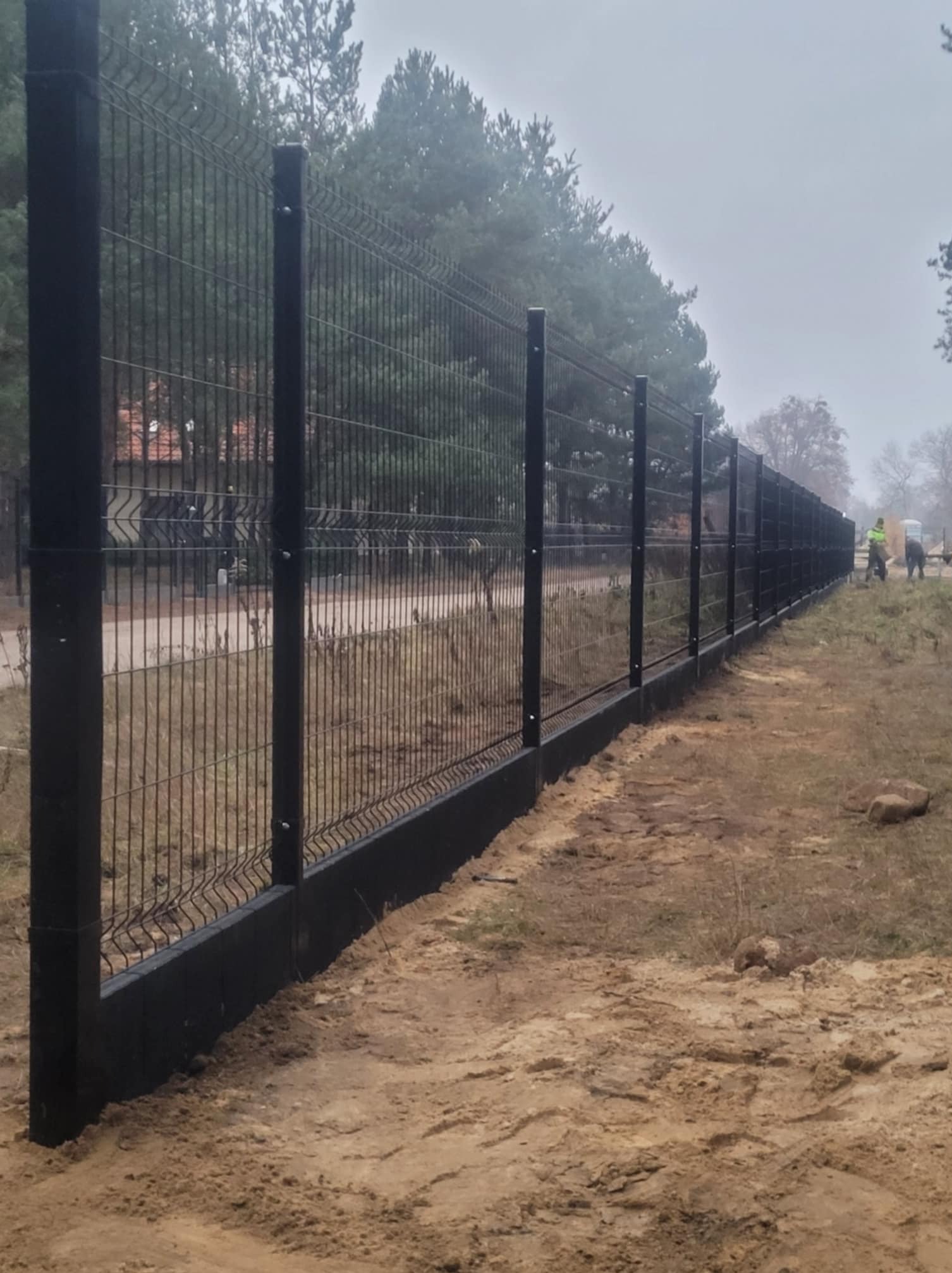 Długi, czarny system ogrodzeniowy z siatki panelowej, z betonową podmurówką, w trakcie montażu w pochmurny dzień; widoczni pracownicy w oddali.