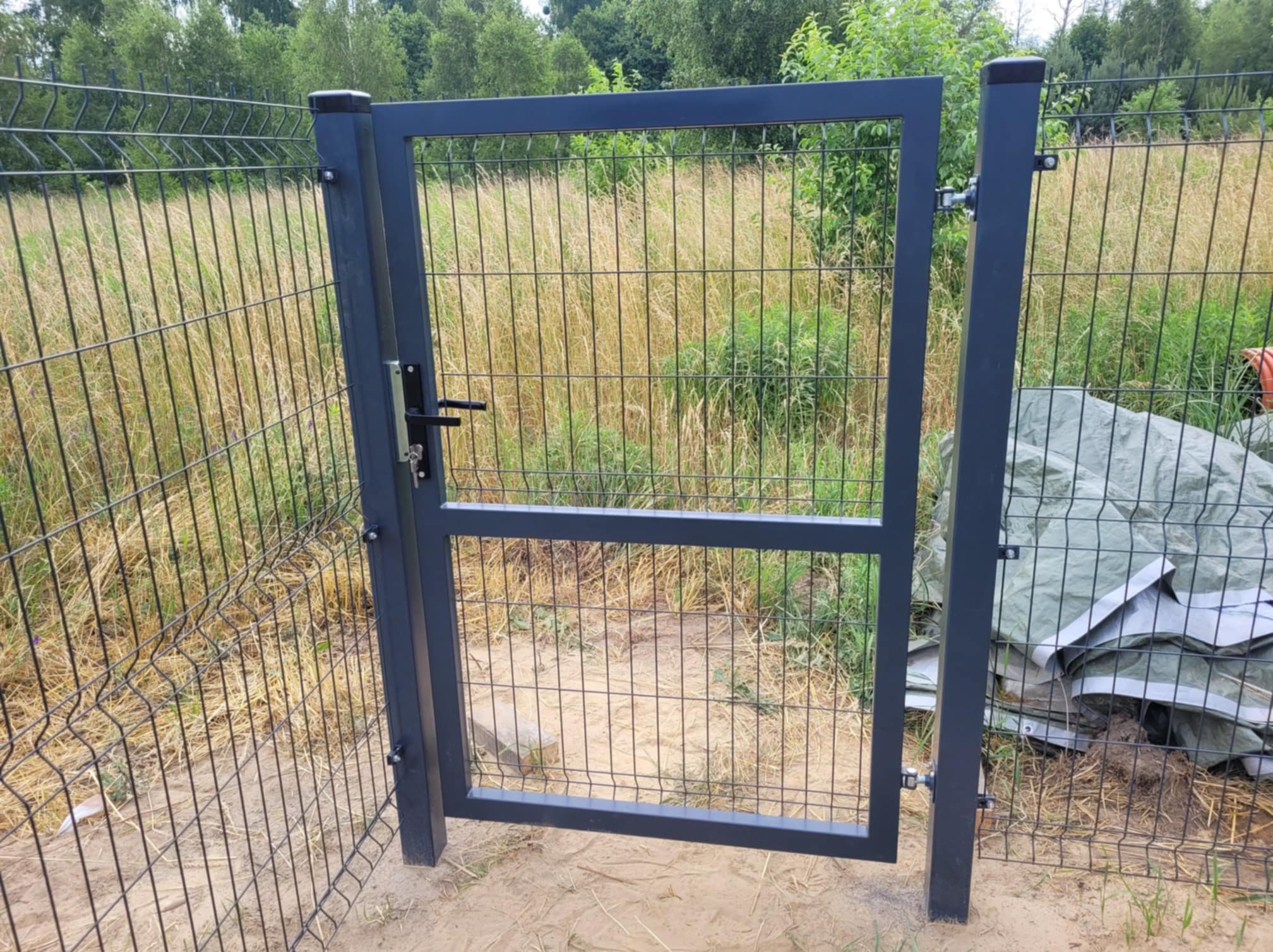 Szara furtka z zamkiem w siatce ogrodzeniowej na tle zarośniętego pola i drzew, widoczne ślady montażu, przy słupku leżą plandeki.
