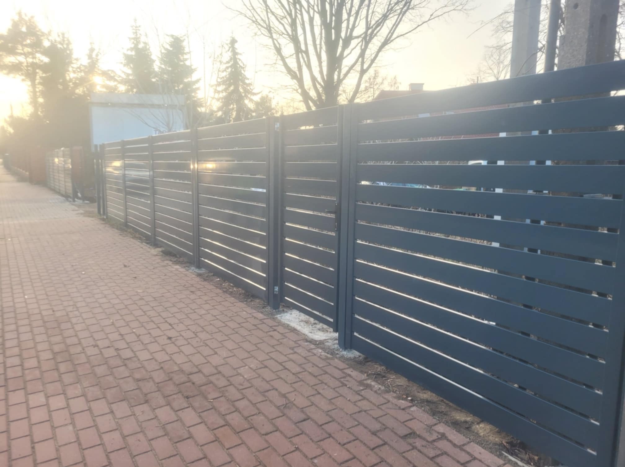 Nowoczesne, poziome ogrodzenie panelowe w kolorze grafitowym, oddzielające chodnik z czerwonej kostki od posesji, w tle drzewa i budynki w porannym słońcu.