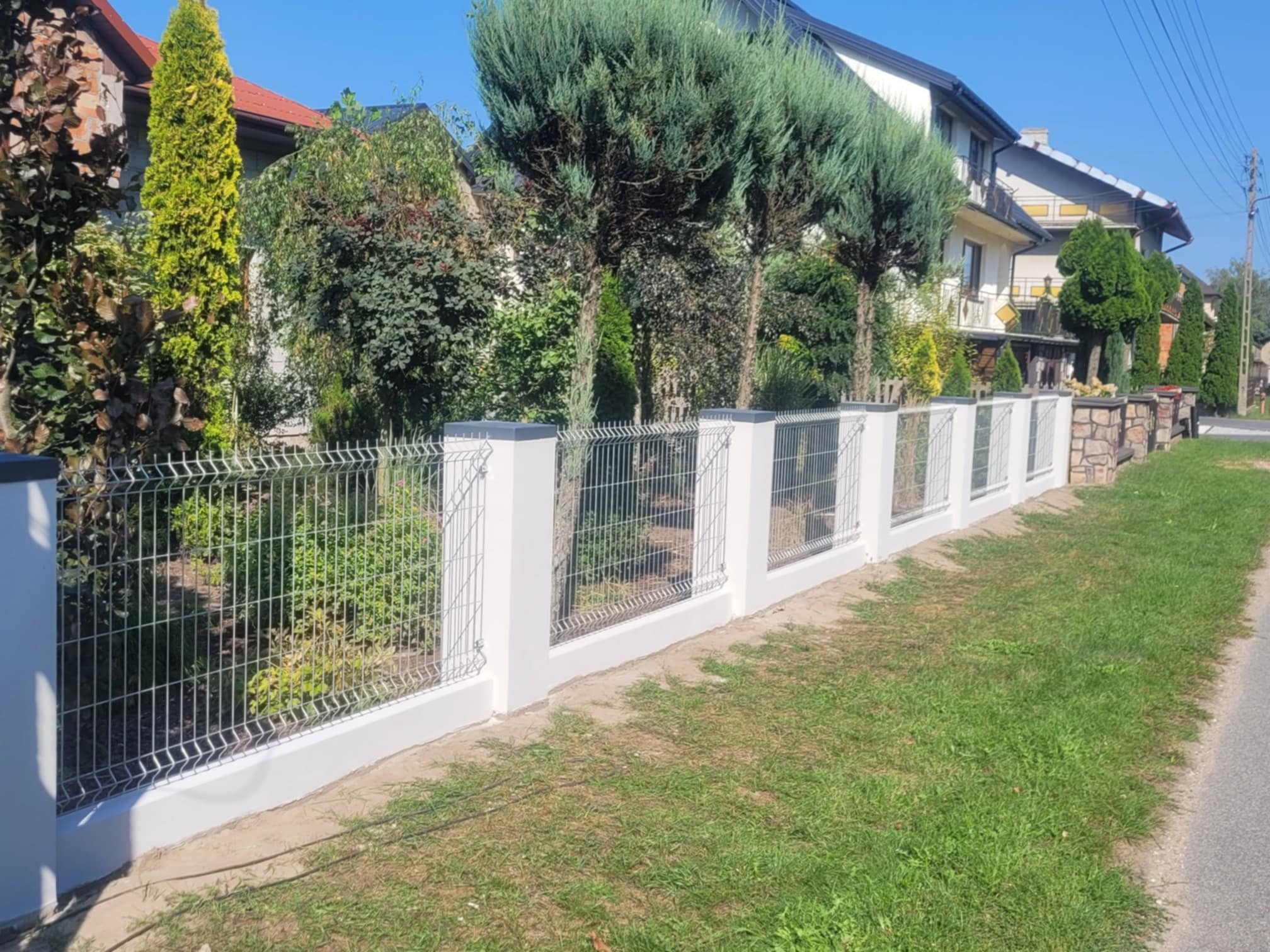 Nowoczesne ogrodzenie z siatki z białymi słupkami, widok z boku, w tle zadbany ogród i domy.