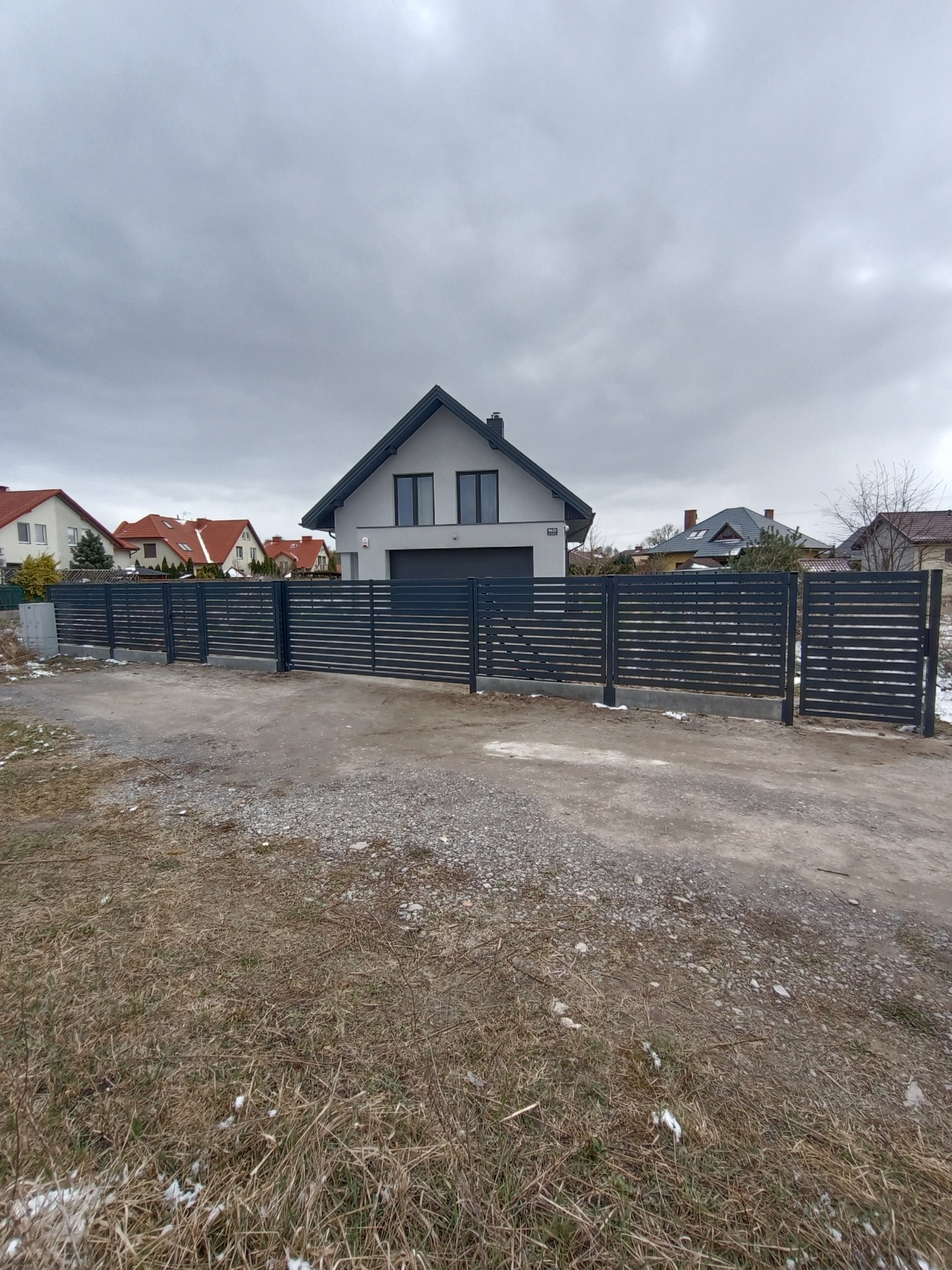 Nowoczesne, poziome przęsła ogrodzeniowe w kolorze grafitowym, z bramą wjazdową, na tle domu z garażem i pochmurnego nieba.