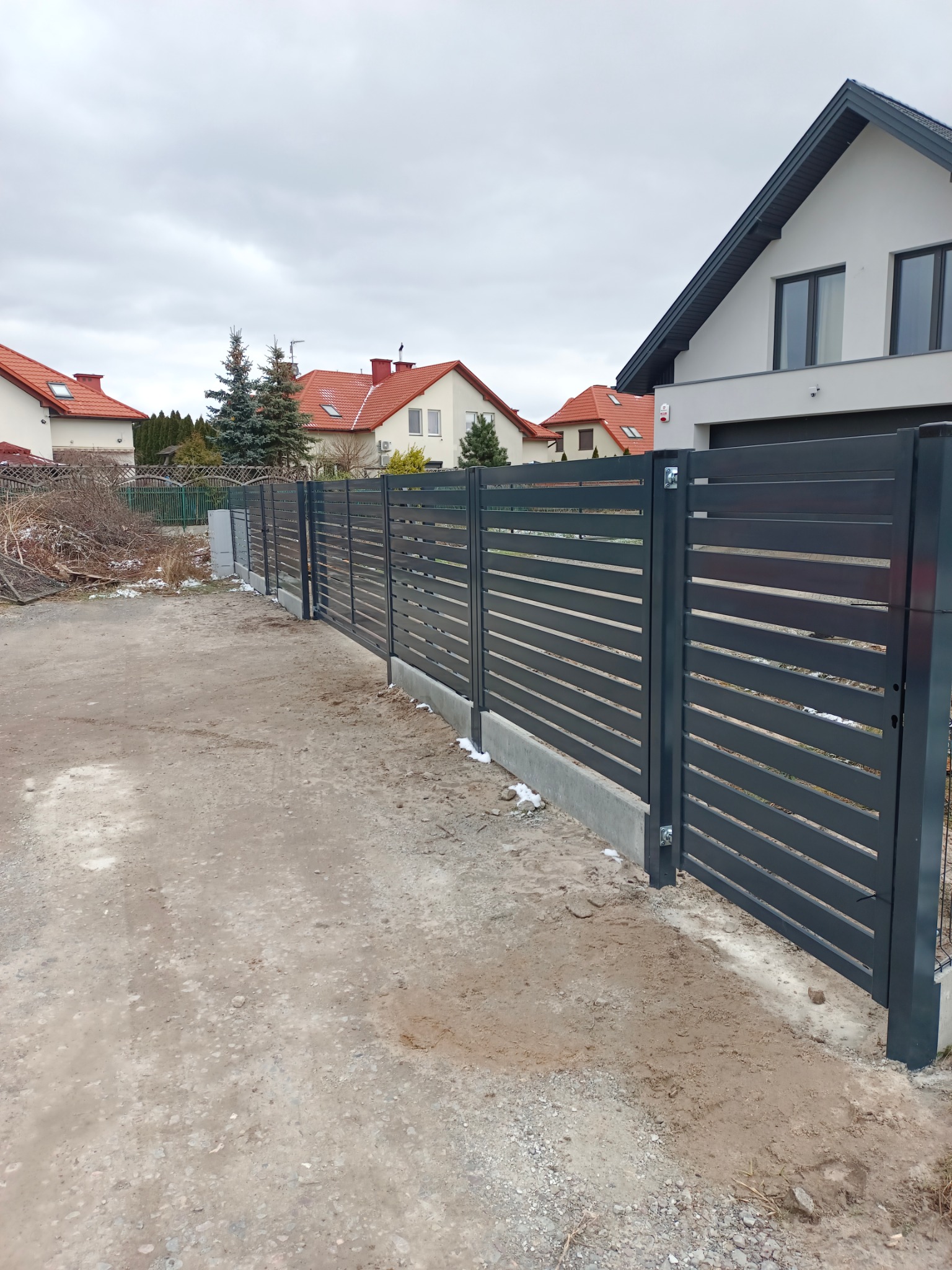 Nowoczesne, poziome, szare przęsła ogrodzeniowe z metalu, osadzone na betonowej podmurówce, przed domem z szarą elewacją w pochmurny dzień.