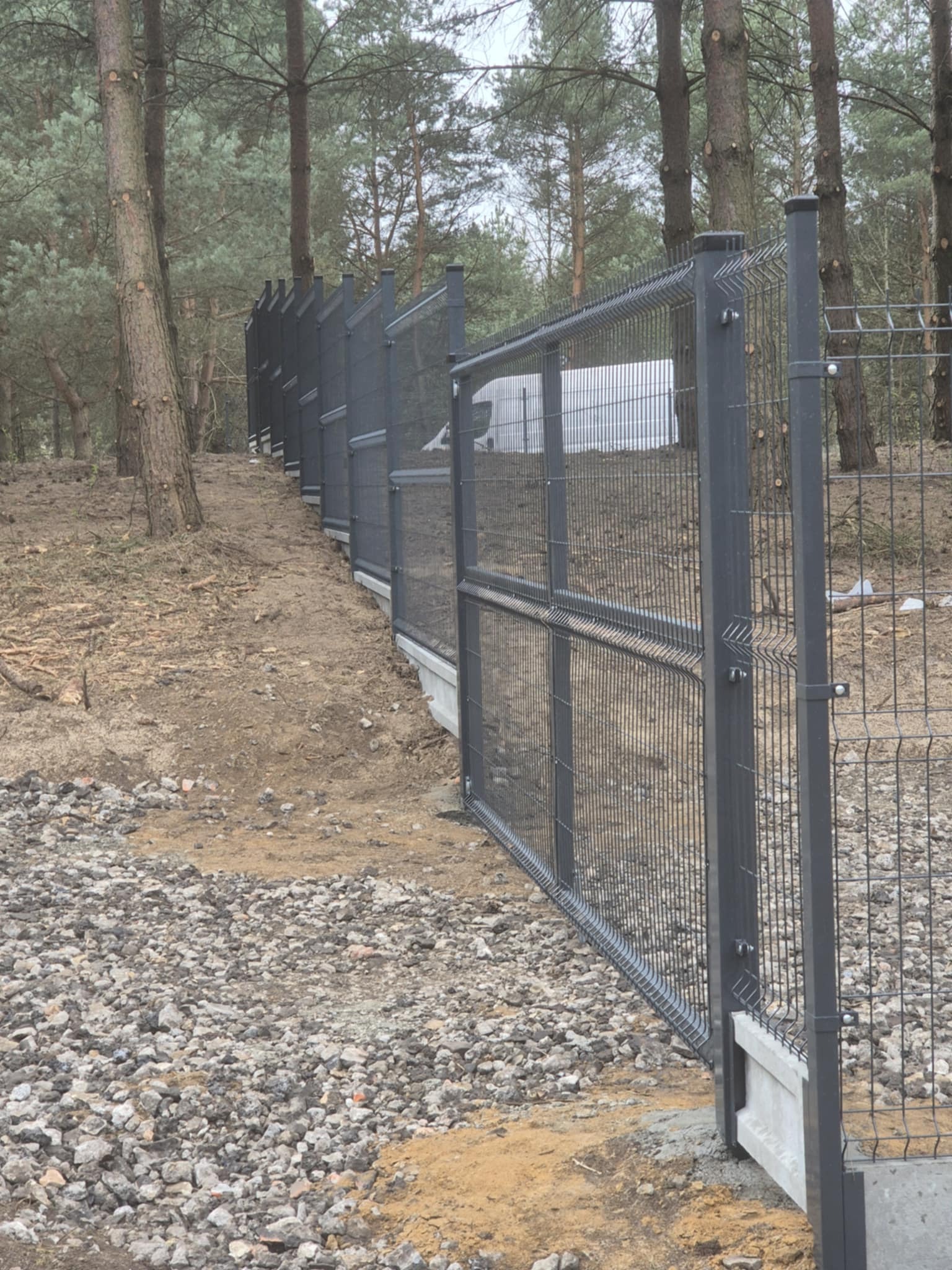 Długi, ciemnoszary płot z paneli z siatki, wsparty na betonowej podmurówce, biegnie wzdłuż leśnego terenu pokrytego kamieniami i igliwiem.
