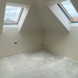 Rach, Ciach, Ciach Dawid Błach Usługi remontowe - Poddasze w trakcie wykańczania, surowe ściany pomalowane na biało, dwa okna dachowe z widokiem na dach sąsiedniego budynku, widoczne przewody elektryczne wystające ze ściany.