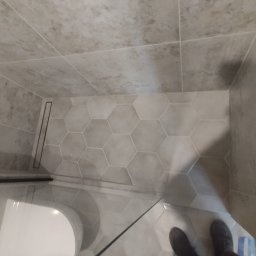 Rach, Ciach, Ciach Dawid Błach Usługi remontowe - Wykończona łazienka z heksagonalną podłogą w odcieniach szarości, liniowy odpływ prysznicowy i częściowo widoczna toaleta oraz fragment butów osoby fotografującej.