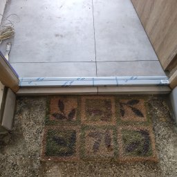 Rach, Ciach, Ciach Dawid Błach Usługi remontowe - Widok progu drzwi wejściowych z nową listwą przypodłogową, częściowo zabezpieczoną folią, położone płytki podłogowe i wycieraczka z motywem roślinnym, widoczny kabel i szpachelka.