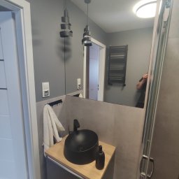 Rach, Ciach, Ciach Dawid Błach Usługi remontowe - Nowoczesna łazienka w szarościach z czarną okrągłą umywalką nablatową, drewnianym blatem i wiszącymi lampami spiralnymi. W lustrze odbicie osoby robiącej zdjęcie.