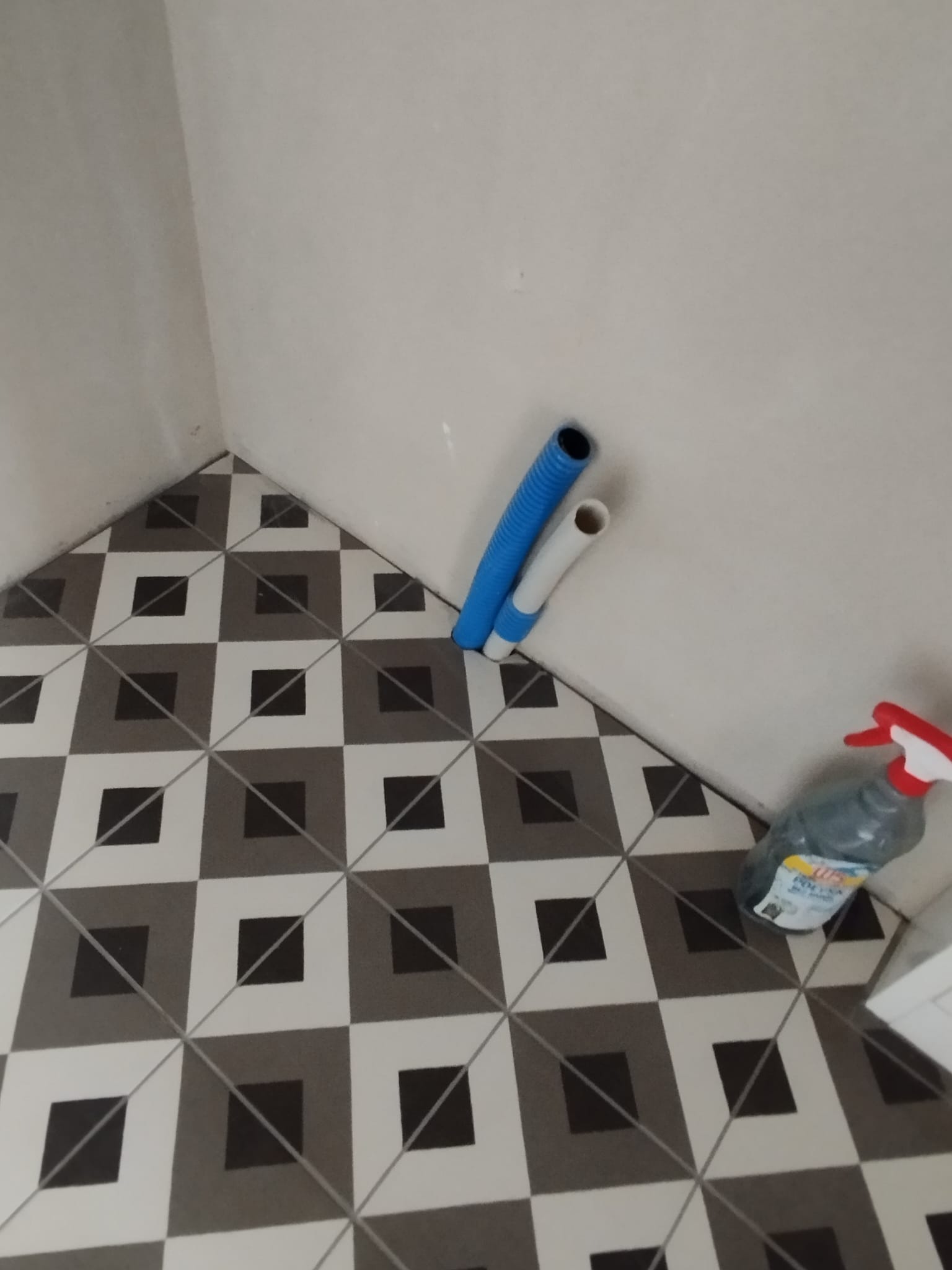 Fragment łazienki po remoncie: podłoga z wzorem geometrycznym, wystające rury instalacyjne oraz butelka środka czyszczącego w rogu.