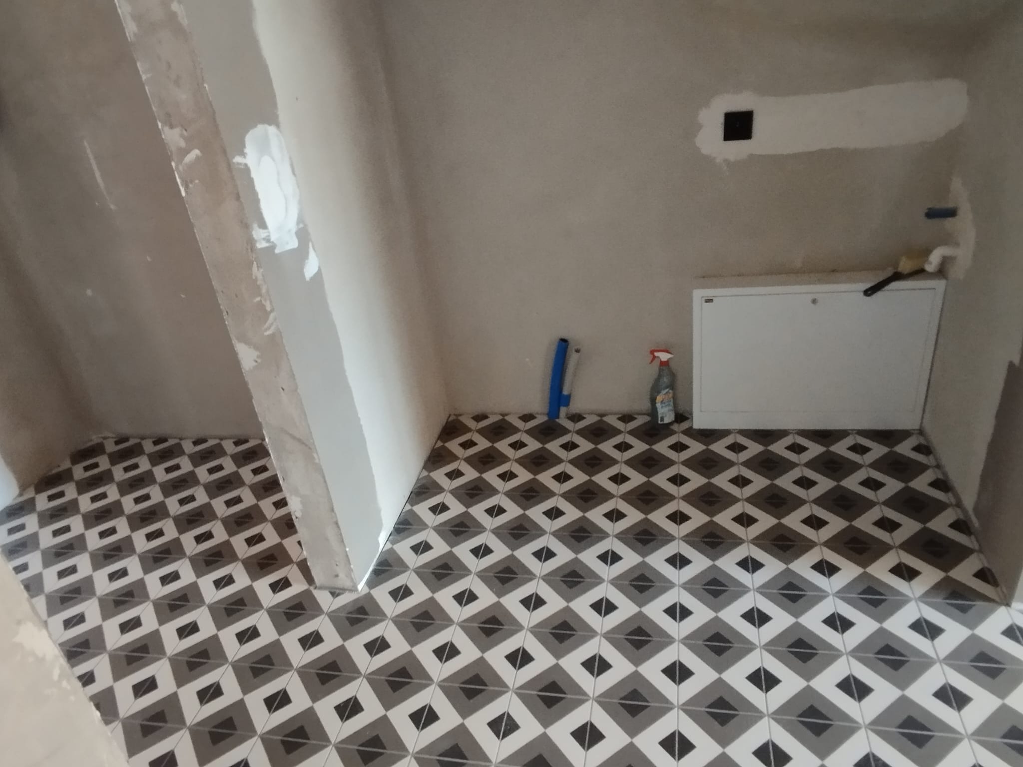 Wnętrze łazienki w trakcie remontu: świeżo położona podłoga z płytek w geometryczny wzór, ściany w surowym tynku, widoczne narzędzia i chemia budowlana, wnęka z pionem kanalizacyjnym.