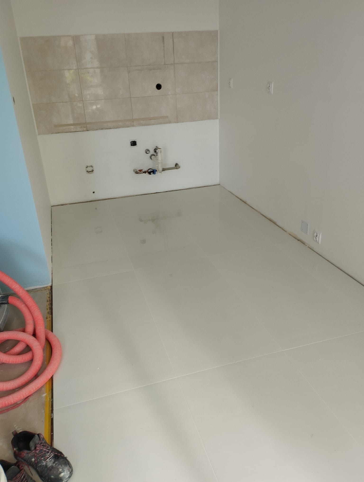 Białe płytki podłogowe w trakcie układania w nowym pomieszczeniu, widoczne przewody instalacyjne i robocze buty w rogu.
