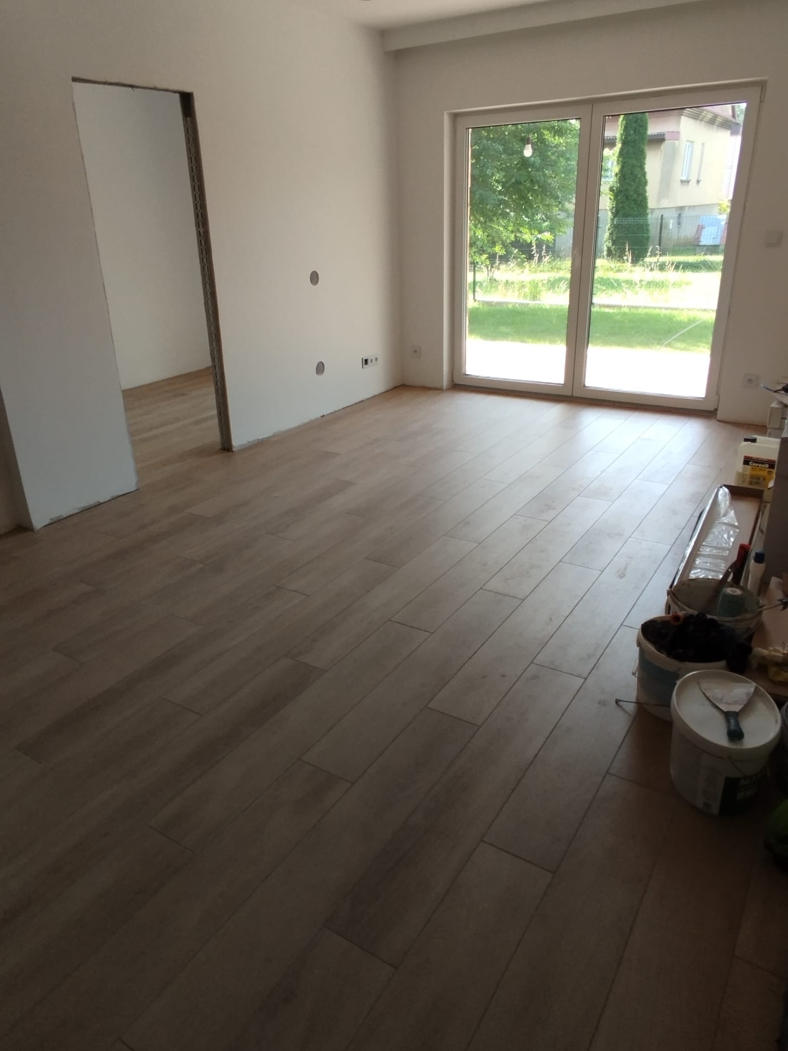 Jasne, drewnopodobne płytki podłogowe ułożone w przestronnym, jasnym pokoju z dużym oknem i przygotowanym wejściem bez drzwi; widoczne wiadra i narzędzia malarskie w rogu.