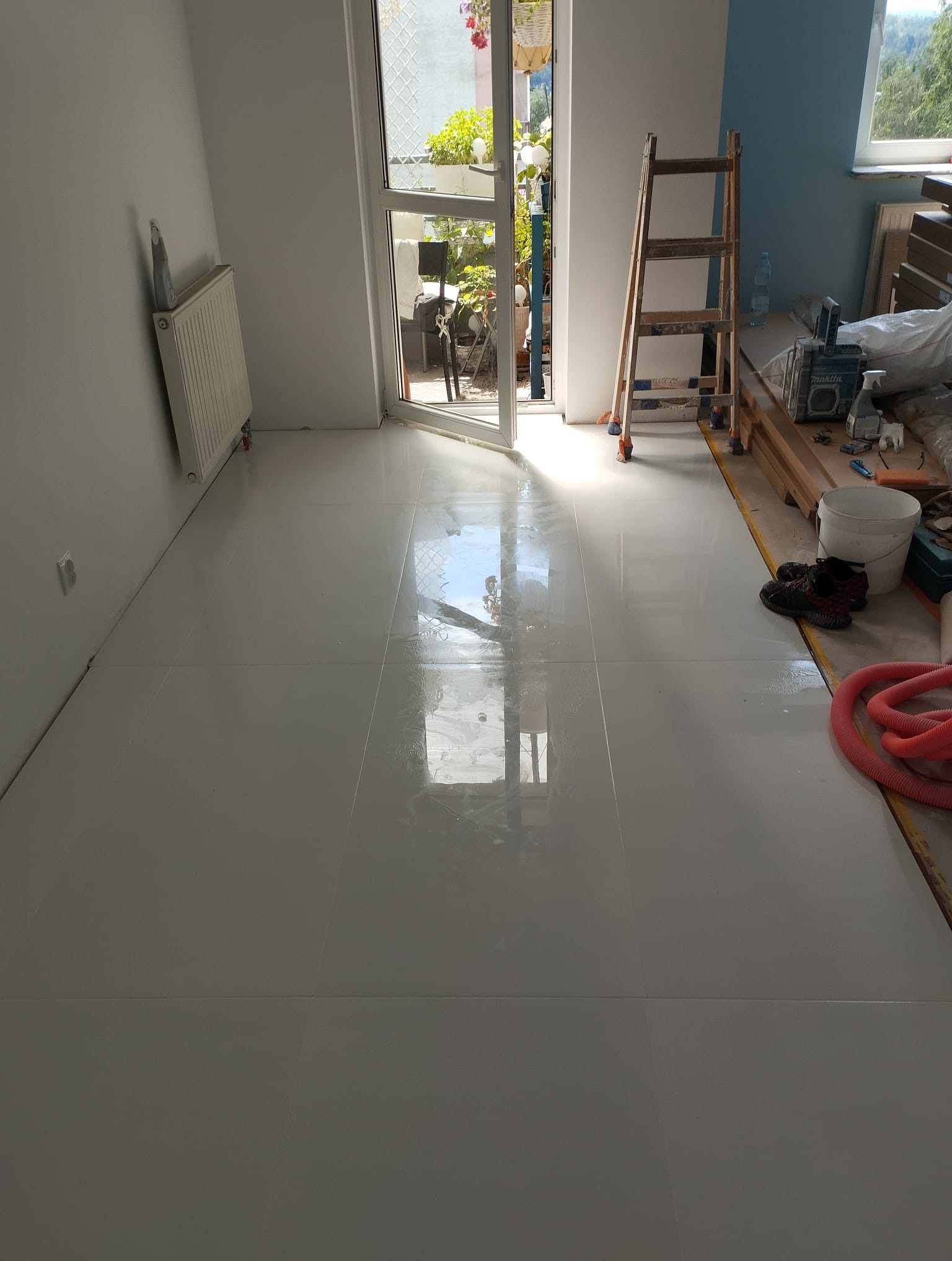 Błyszcząca, jasna podłoga z płytek po ułożeniu, widoczne narzędzia i drabina w tle, otwarte drzwi balkonowe wpuszczające światło.