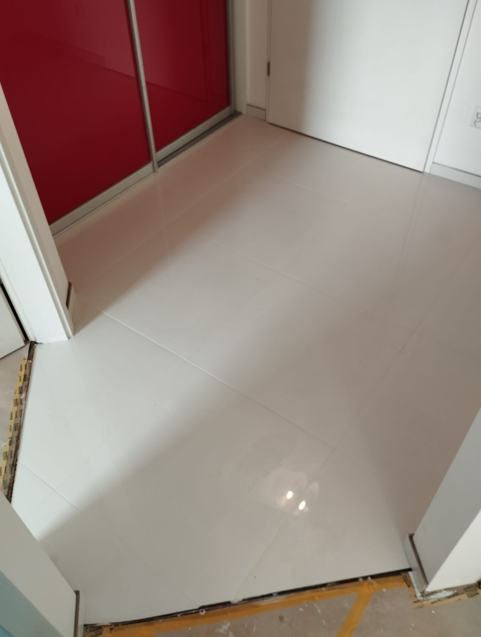 Świeżo położona, błyszcząca, jasna terakota w przedpokoju, widoczne niedokończone listwy przypodłogowe i fragment czerwonej szafy przesuwnej.