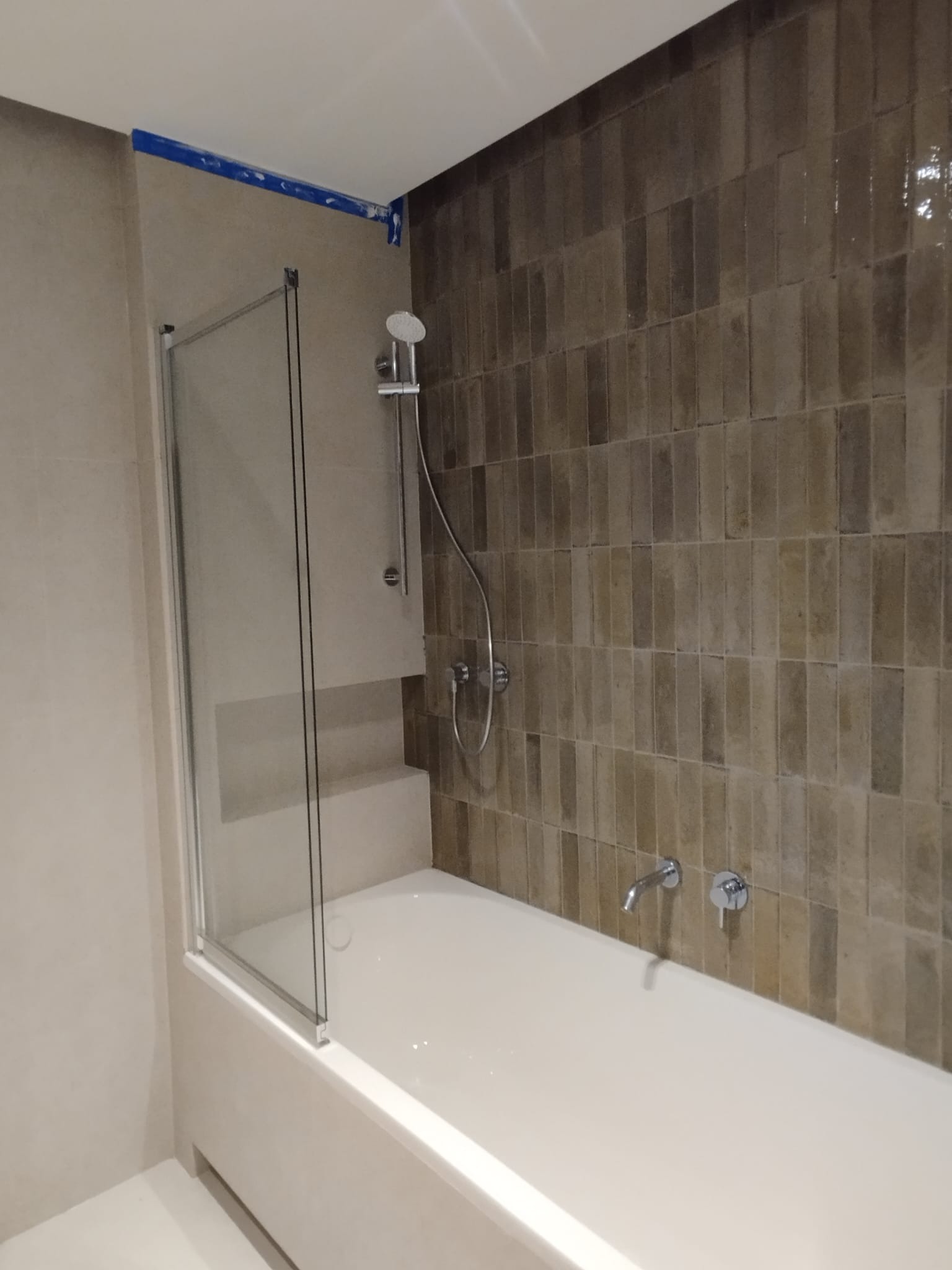 Wykończona łazienka z wanną z parawanem, brązowe płytki ułożone pionowo, bateria wannowa zamontowana na ścianie, widoczna taśma malarska na suficie.