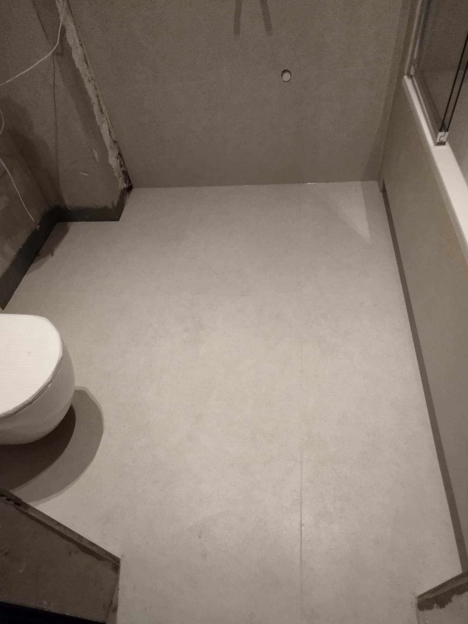 Wnętrze łazienki w trakcie prac wykończeniowych, widoczna surowa ściana pod skosem, częściowo ułożona jasna podłoga z płytek i fragment toalety.