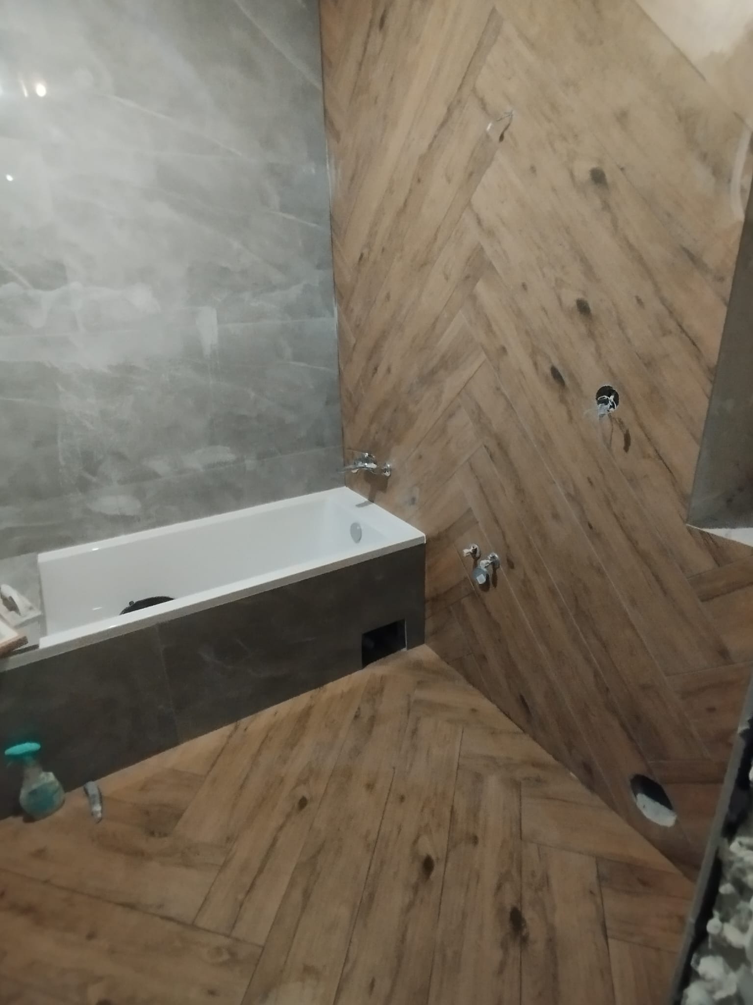 Łazienka w trakcie remontu: widoczna wanna z szarą obudową, fragment ściany wyłożonej szarymi płytkami imitującymi marmur oraz ściana i podłoga wyłożone drewnopodobnymi płytkami w jodełkę,...