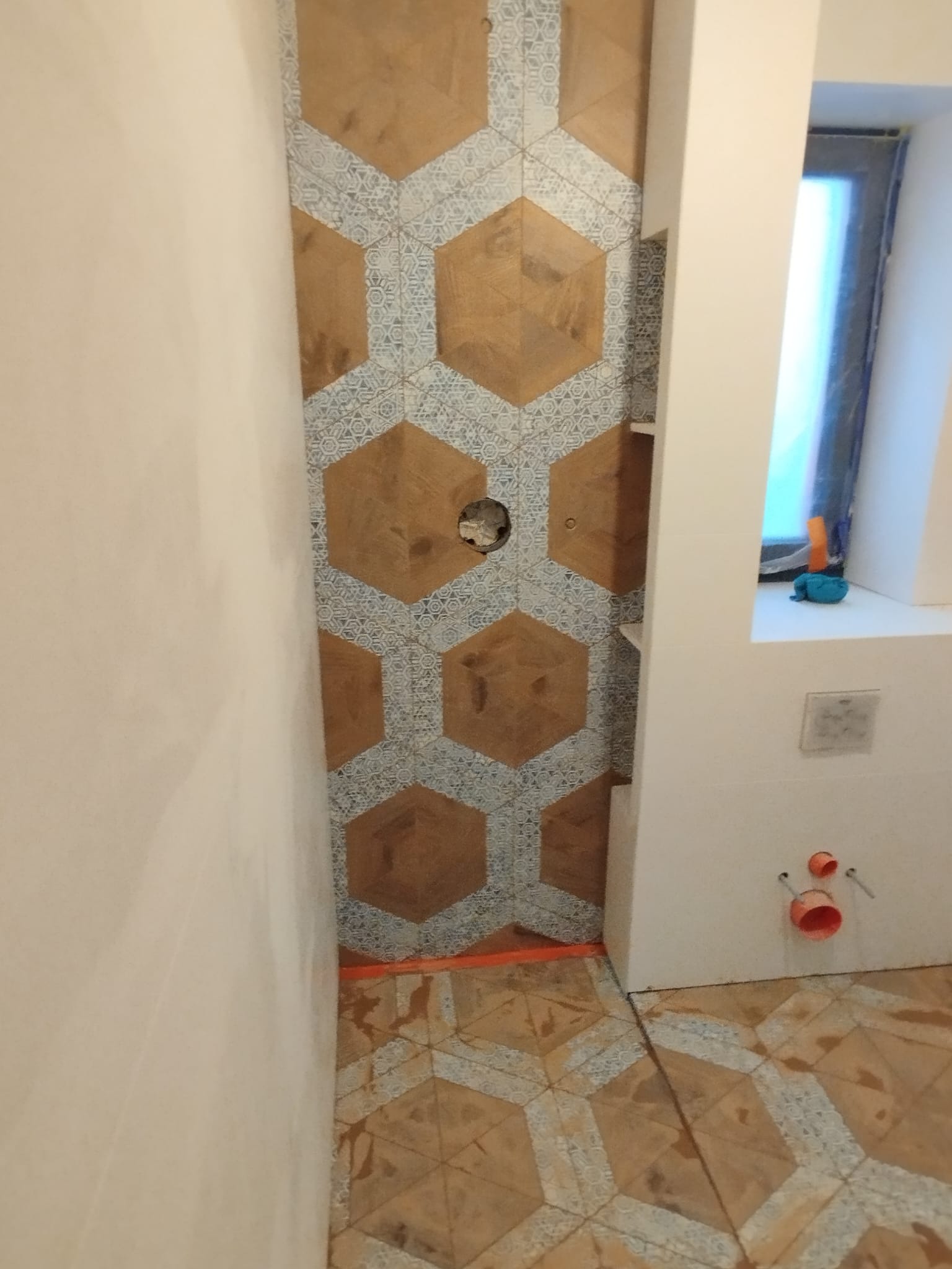 Ściana i podłoga w łazience wyłożone heksagonalnymi płytkami w odcieniach brązu i szarości, z widocznymi elementami instalacji hydraulicznej i elektrycznej przed montażem armatury.