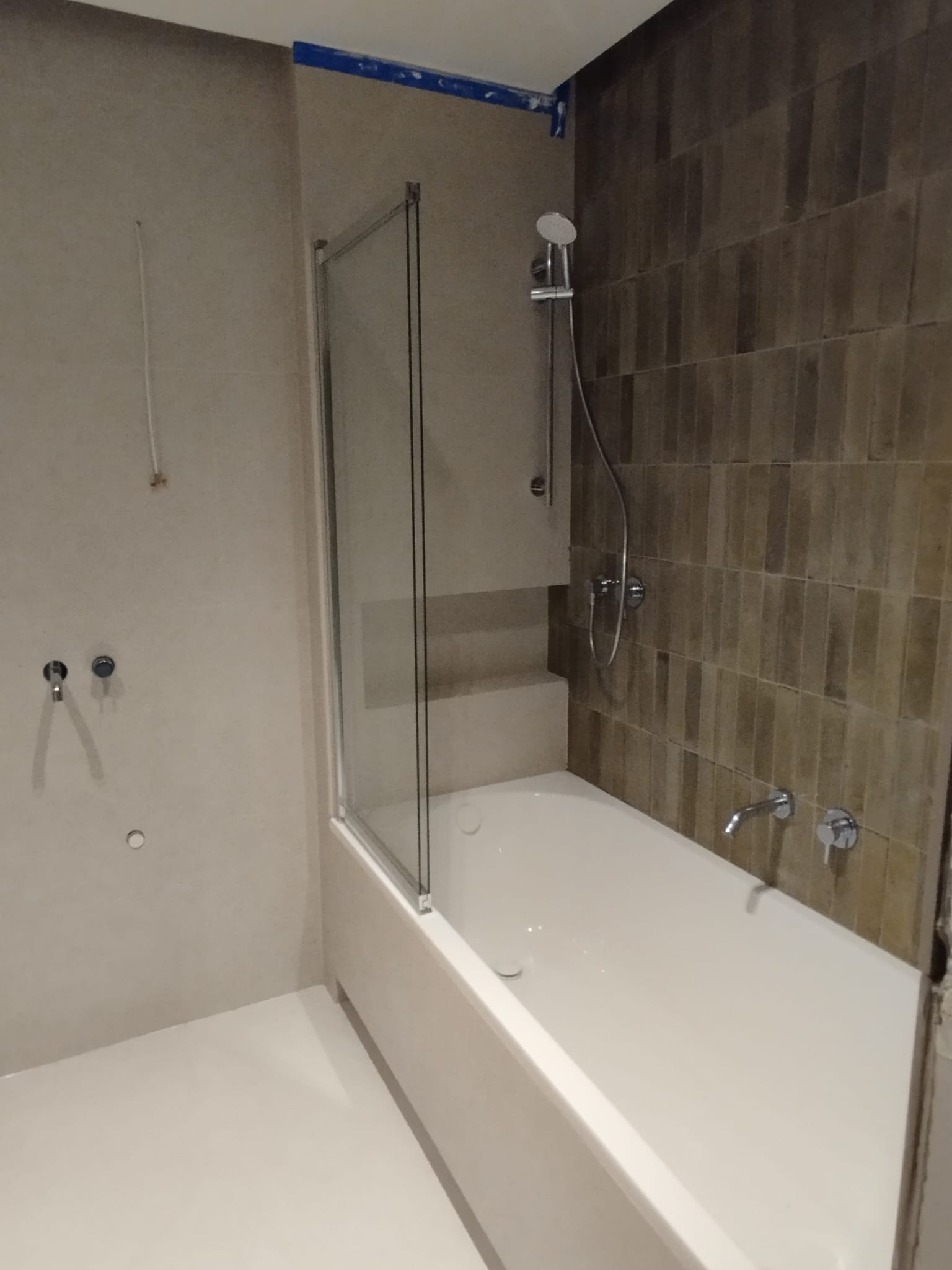 Nowoczesna łazienka z beżowymi ścianami, kabiną prysznicową z przezroczystego szkła i chromowaną armaturą; widoczne ślady taśmy malarskiej na suficie.