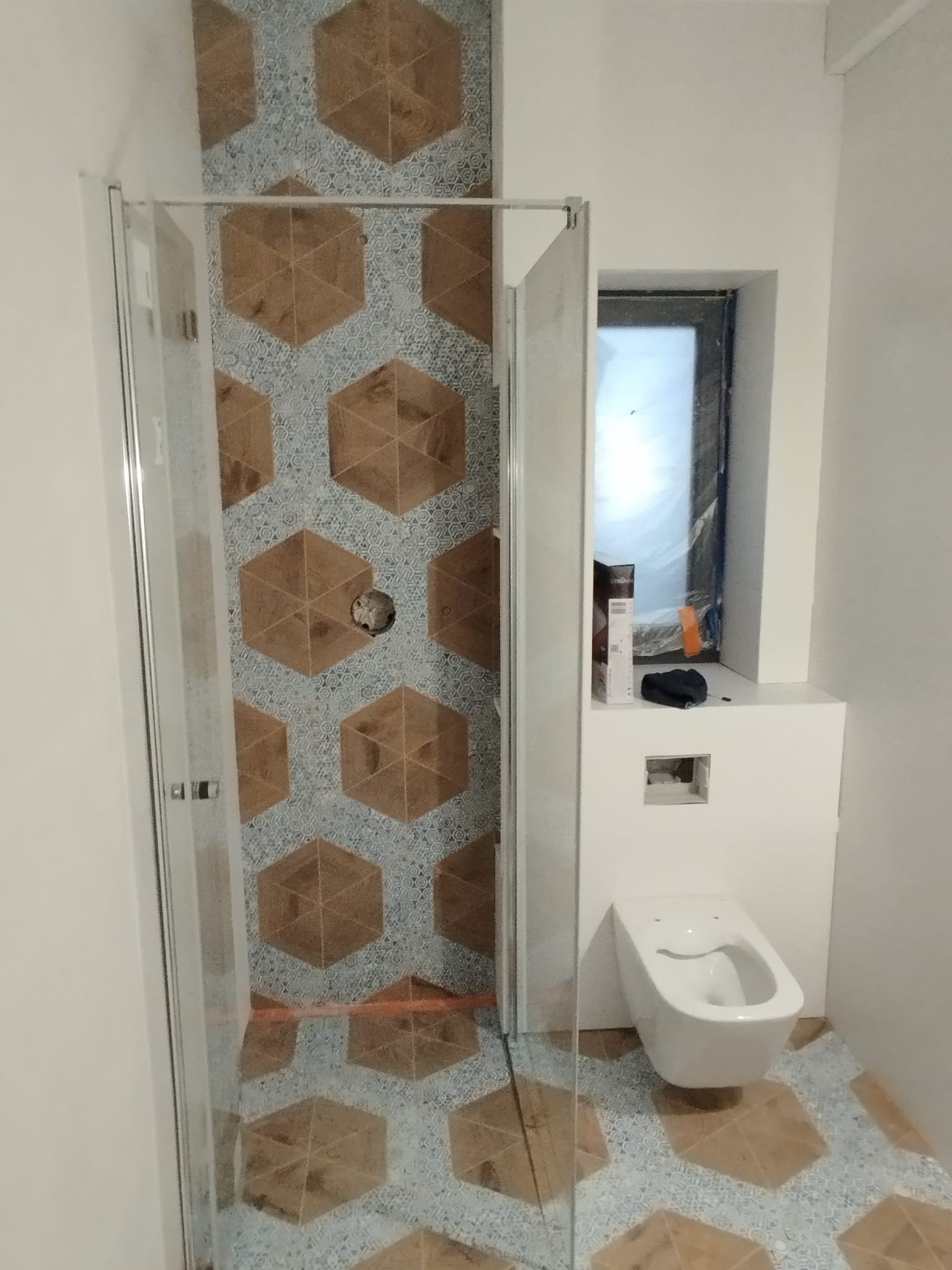 Nowoczesna łazienka w trakcie wykańczania, widoczna kabina prysznicowa z przezroczystymi drzwiami, podwieszana toaleta oraz heksagonalne płytki w odcieniach brązu i błękitu na ścianach i podłodze...