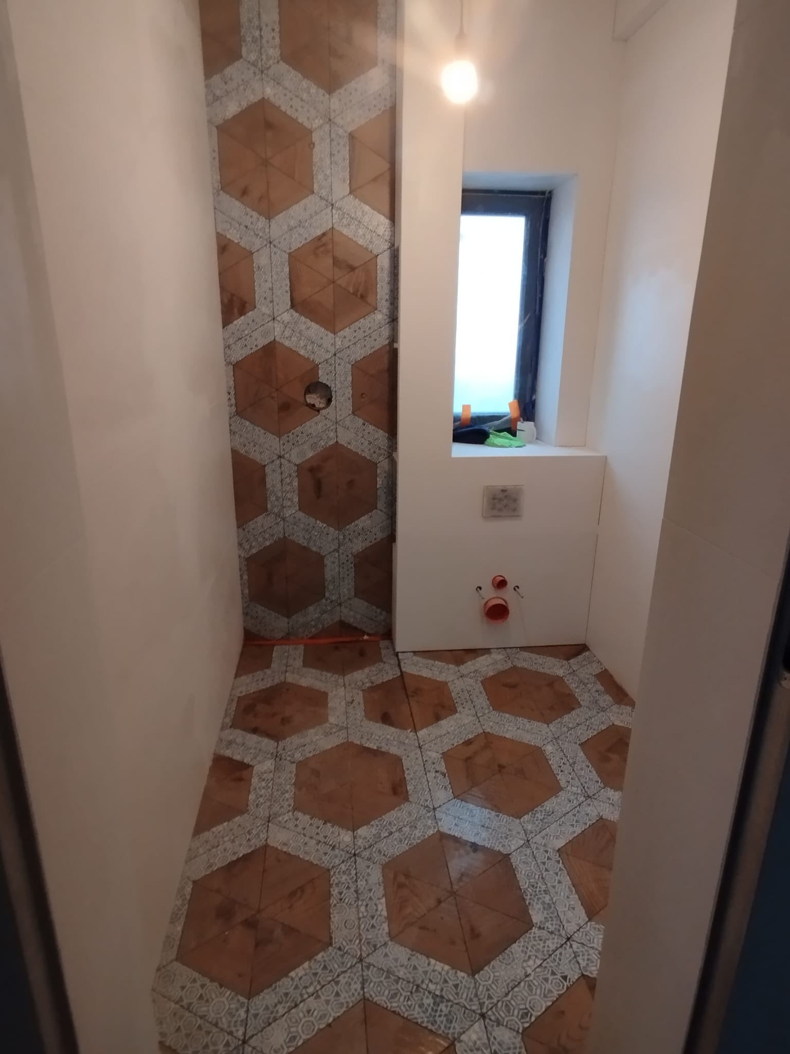 Łazienka w trakcie prac wykończeniowych: ułożone heksagonalne płytki podłogowe i ścienne w odcieniach brązu i szarości, widoczne rury instalacyjne, okno z akcesoriami i wisząca żarówka.