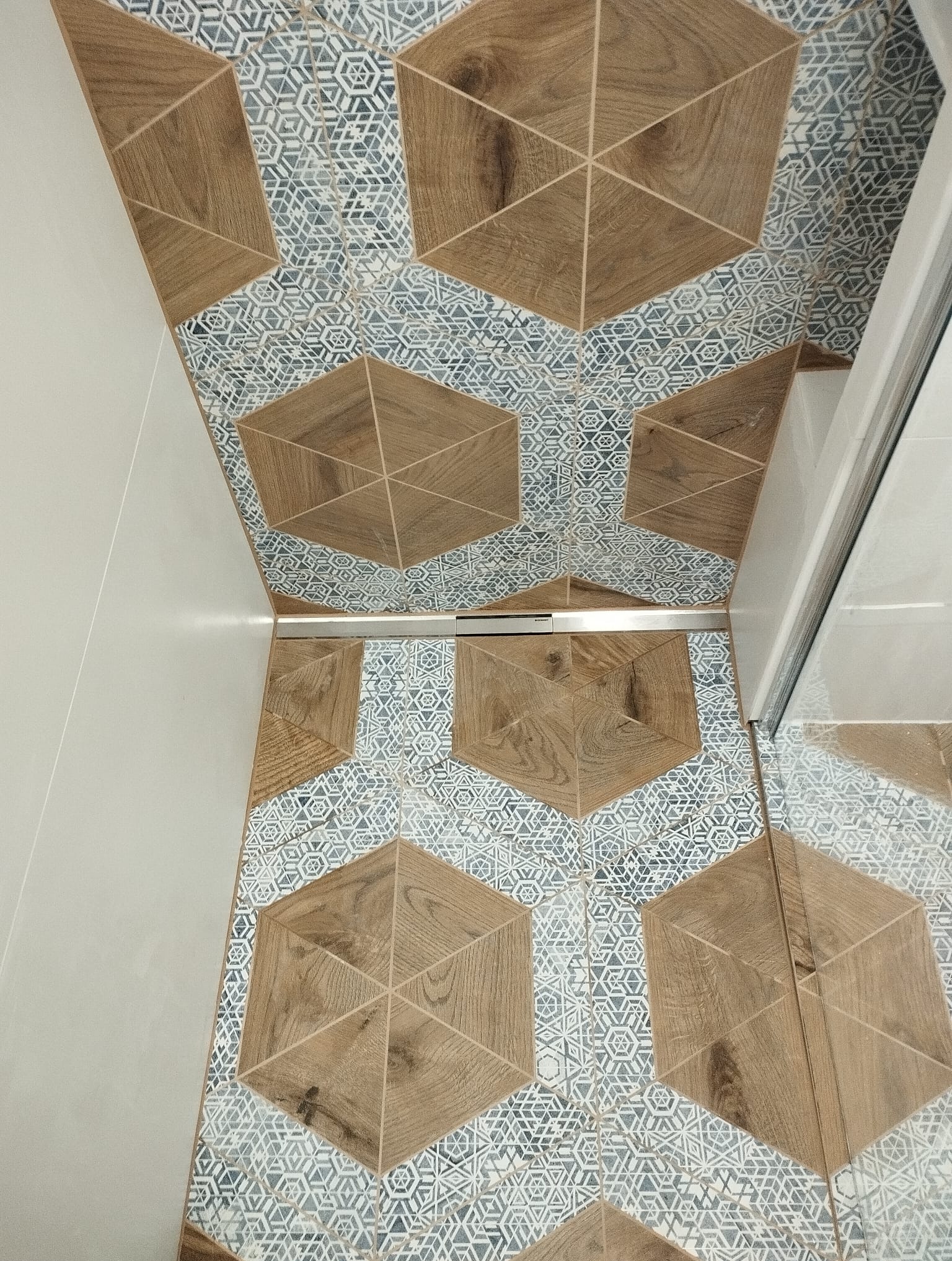 Wzór podłogi łazienkowej z płytek ceramicznych w kształcie heksagonów z motywem drewna i geometrycznymi wzorami w odcieniach szarości, widoczny odpływ liniowy i fragment kabiny prysznicowej.