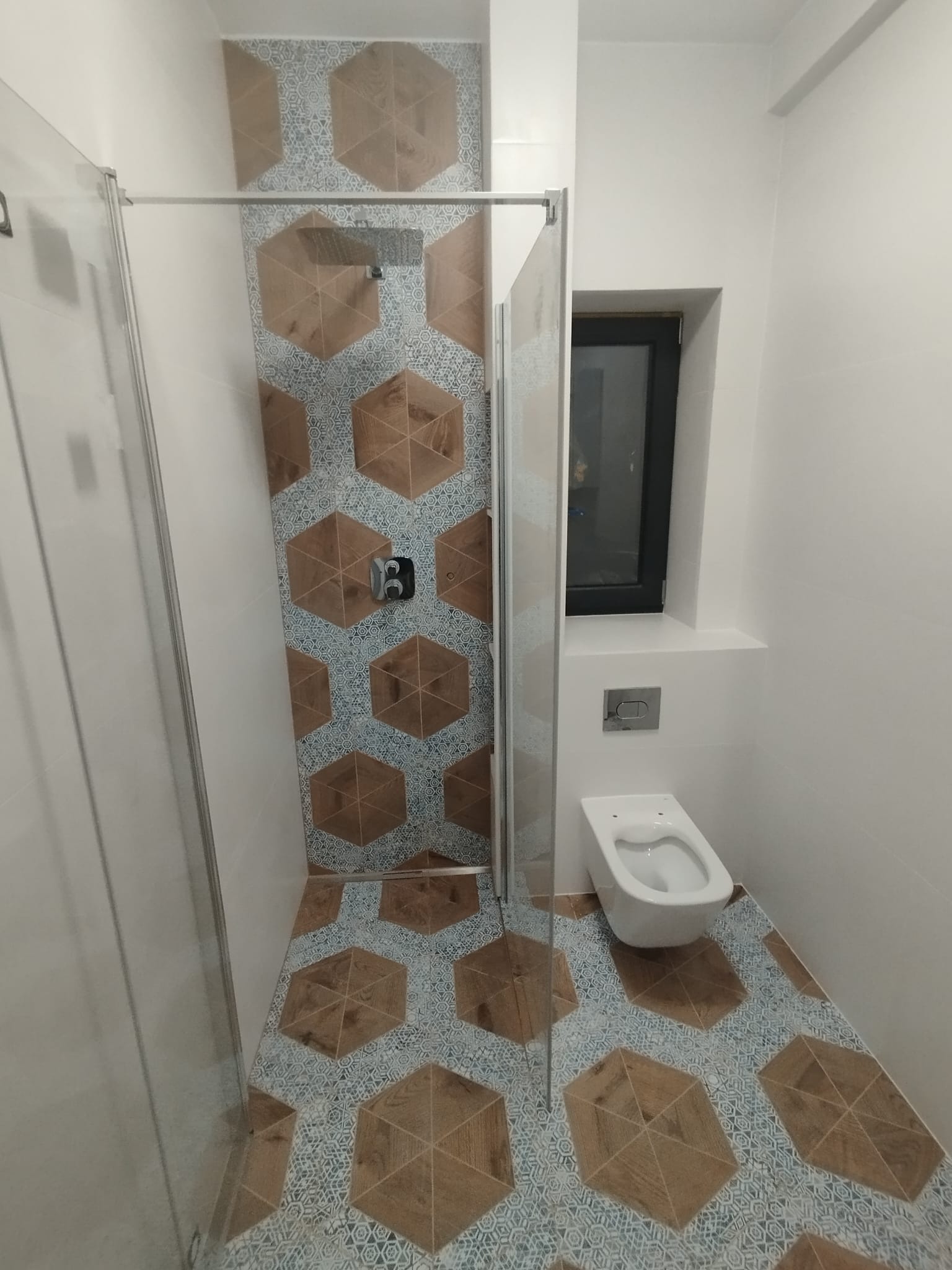 Nowoczesna łazienka z kabiną prysznicową typu walk-in, wiszącą miską WC i geometrycznymi płytkami w odcieniach brązu i błękitu.