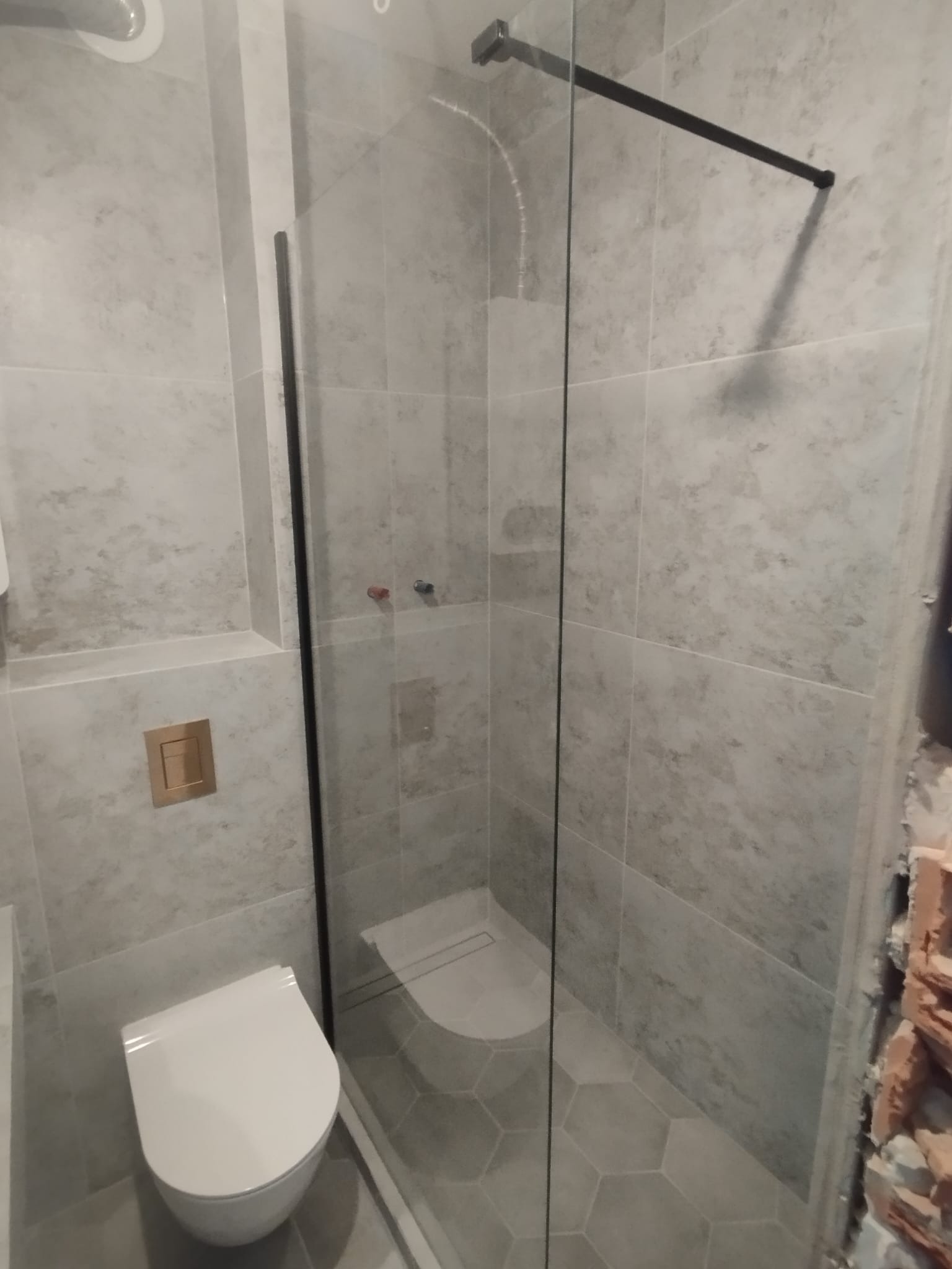 Nowoczesna łazienka z kabiną prysznicową typu walk-in z czarnymi profilami, podwieszaną toaletą z przyciskiem w kolorze mosiądzu i heksagonalnymi płytkami podłogowymi w odcieniach szarości...