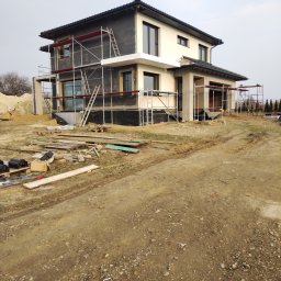 APG CONSTRUCTION SPÓŁKA Z OGRANICZONĄ ODPOWIEDZIALNOŚCIĄ - Dwukondygnacyjny dom w trakcie ocieplania elewacji, z widocznymi rusztowaniami i materiałami budowlanymi na posesji. Część budynku otynkowana na kremowo.