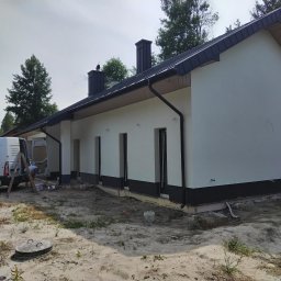 APG CONSTRUCTION SPÓŁKA Z OGRANICZONĄ ODPOWIEDZIALNOŚCIĄ - Elewacja domu jednorodzinnego w trakcie prac wykończeniowych. Widoczny fragment ocieplenia fundamentów, biała elewacja i pracownik przy samochodzie dostawczym.