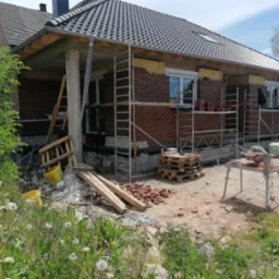 Budowa domu z cegły: ściany w trakcie wznoszenia, widoczne rusztowania, izolacja i narzędzia. Częściowo wykończona elewacja, niekompletny stan budynku.