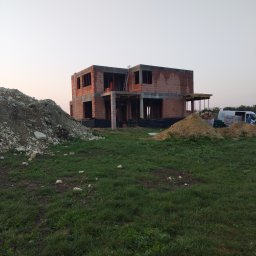 APG CONSTRUCTION SPÓŁKA Z OGRANICZONĄ ODPOWIEDZIALNOŚCIĄ - Surowy stan dwukondygnacyjnego domu z cegły, z widocznym szkieletem i stertami ziemi, na tle zielonej trawy i bladego nieba o zmierzchu. Widoczny częściowo samochód dostawczy.