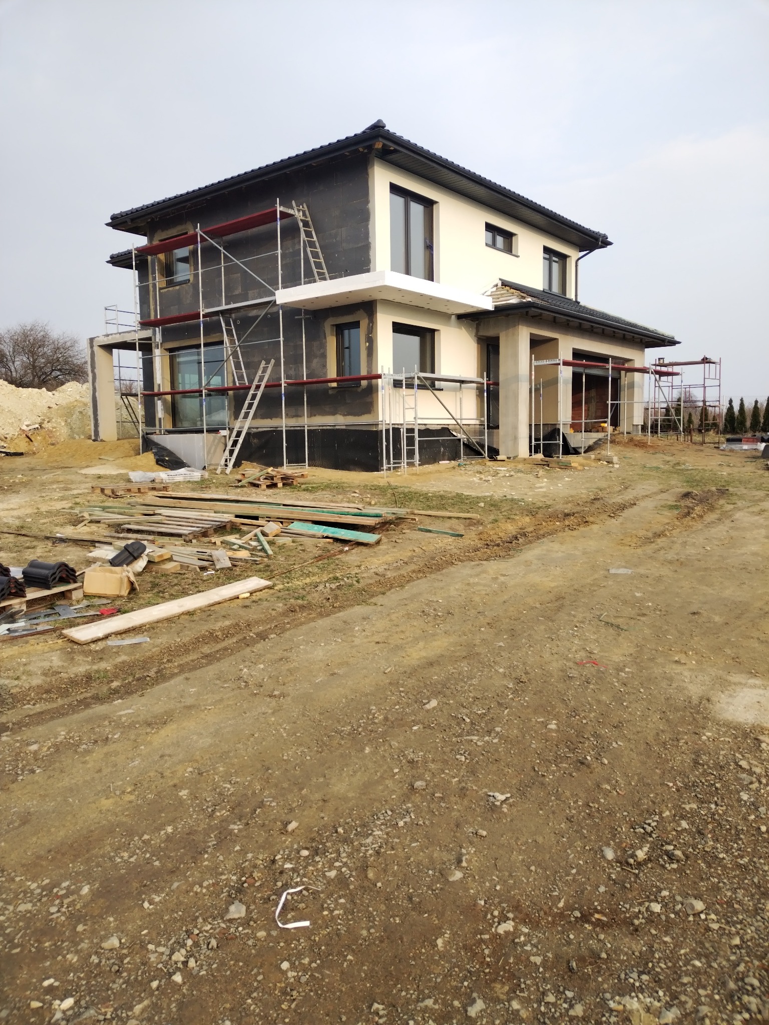 Dwukondygnacyjny dom w trakcie ocieplania elewacji, z widocznymi rusztowaniami i materiałami budowlanymi na posesji. Część budynku otynkowana na kremowo.