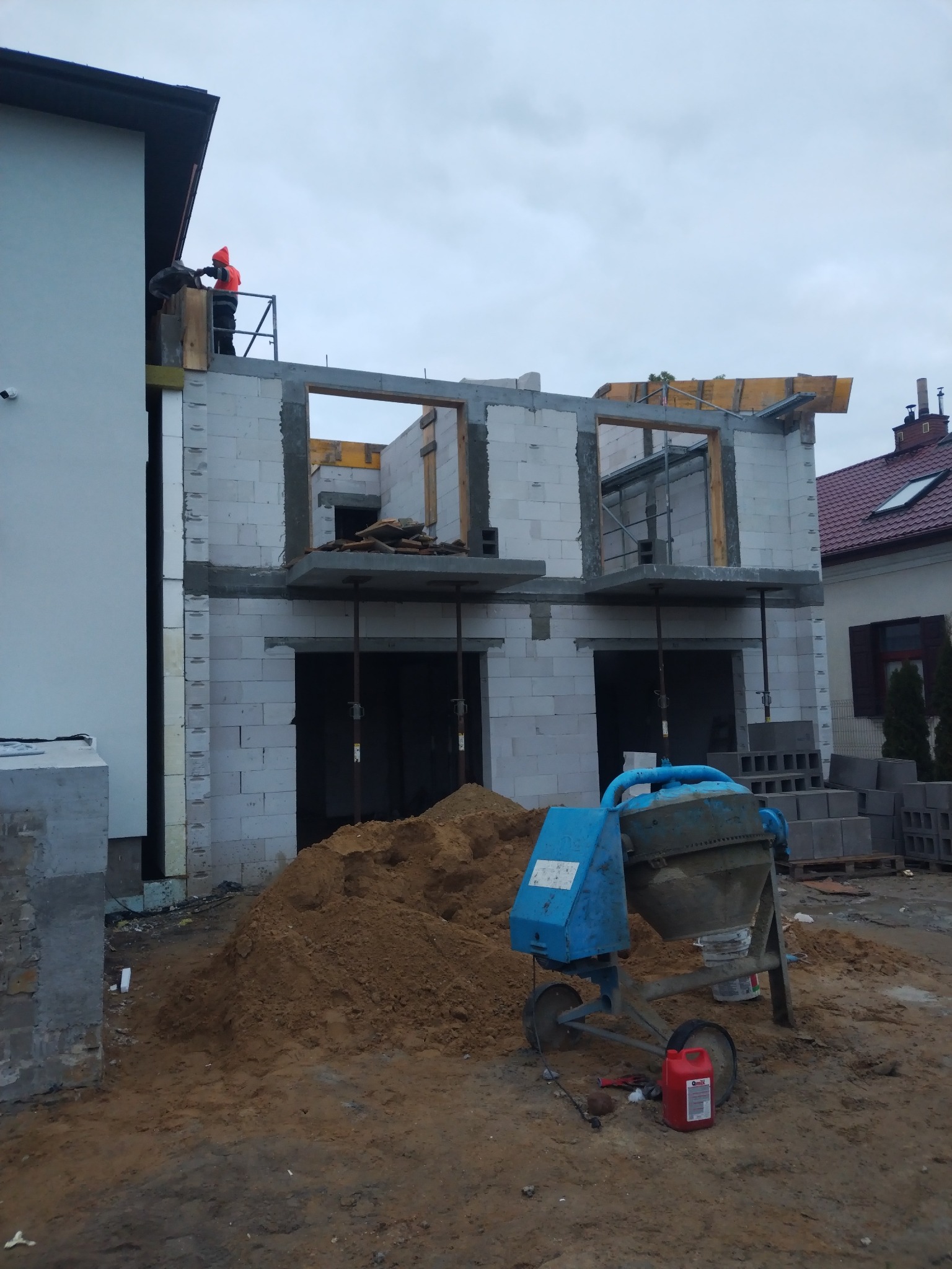 Budowa domu z betonu komórkowego w trakcie realizacji, widoczna betoniarka i pracownik na rusztowaniu. Surowy stan budynku, okna bez szyb.